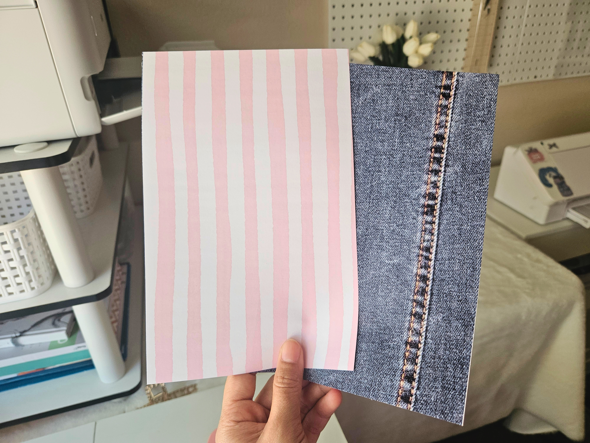 Hand holding a notebook with pink striped cover and denim side, office setting in the background