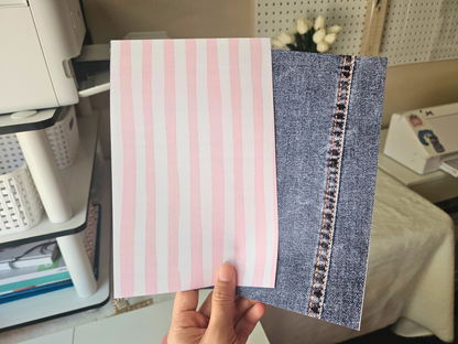 Hand holding a notebook with pink striped cover and denim side, office setting in the background