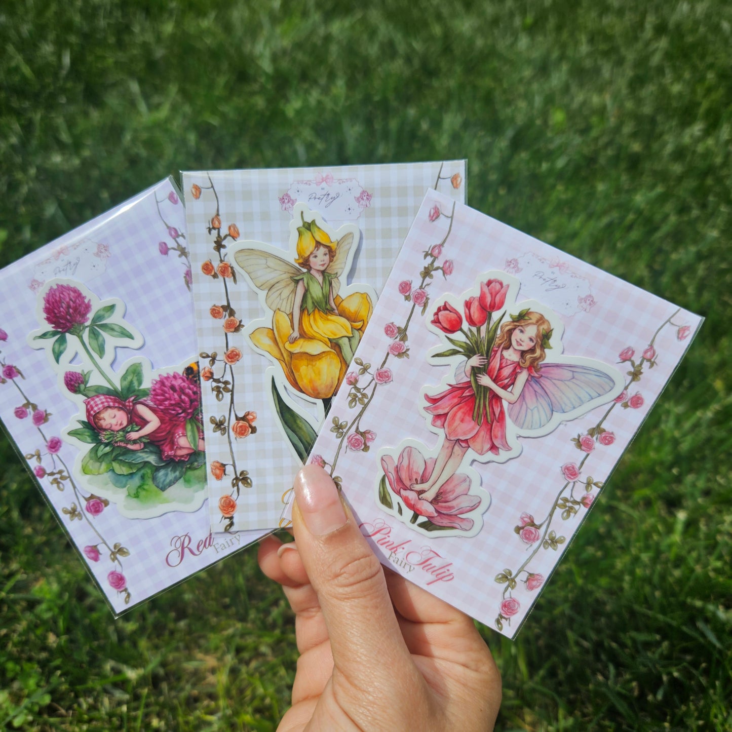 Hand holding three colorful fairy cards against a grassy background